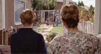Movie still from “Life Is Sweet” (1990), directed by Mike Leigh – Two women watching a man perform a show in a backyard; Wide shot, Over the shoulder angle