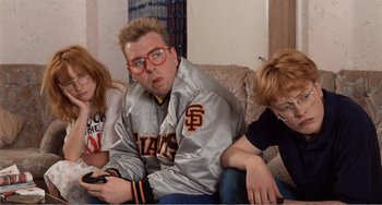 Movie still from “Life Is Sweet” (1990), directed by Mike Leigh – A man sitting on a couch next to two other people holding remotes; Medium shot, Low angle