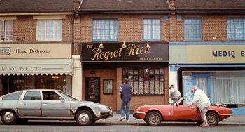 Movie still from “Life Is Sweet” (1990), directed by Mike Leigh – A man standing in front of a restaurant next to two cars; Wide shot, High angle