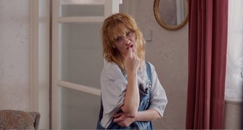 Movie still from “Life Is Sweet” (1990), directed by Mike Leigh – A woman in a white shirt and blue overalls is posing for a picture; Medium shot, Over the shoulder angle