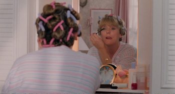 Movie still from “Life Is Sweet” (1990), directed by Mike Leigh – A woman in curlers putting makeup on in front of a mirror; Medium shot, Over the shoulder angle