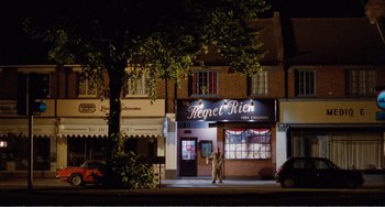 Movie still from “Life Is Sweet” (1990), directed by Mike Leigh – A person standing in front of a store at night; Extreme Wide shot, High angle