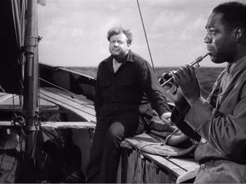 Movie still from “Lifeboat” (1944), directed by Alfred Hitchcock – A man sitting on the side of a boat next to a man playing an instrument; Medium shot, High angle