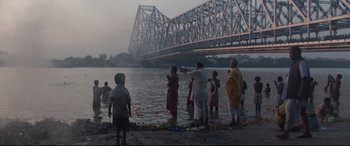 Movie still from “Lion” (2016), directed by Garth Davis – A group of people standing in the water near a bridge; Extreme Wide shot, Low angle