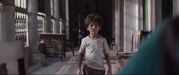 Movie still from “Lion” (2016), directed by Garth Davis – A young boy standing in a room next to a table; Medium shot, Over the shoulder angle