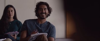 Movie still from “Lion” (2016), directed by Garth Davis – A man sitting on the ground holding a notebook; Medium shot, Over the shoulder angle