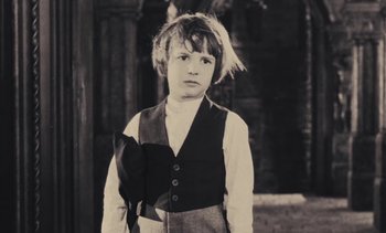 Movie still from “Lions Love (... and Lies)” (1969), directed by Agnès Varda – A young boy wearing a vest standing in front of a building; Medium shot, High angle