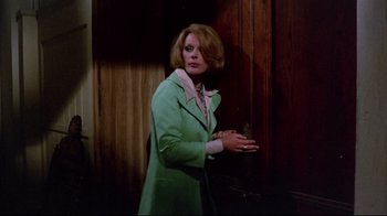 Movie still from “Lisa and the Devil” (1973), directed by Mario Bava – A woman wearing a green coat standing next to a wooden wall; Medium shot, Over the shoulder angle