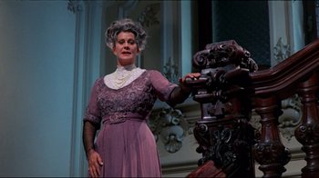 Movie still from “Lisa and the Devil” (1973), directed by Mario Bava – An older woman in a purple dress stands in front of an ornate wooden table; Medium shot, Low angle