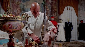 Movie still from “Lisa and the Devil” (1973), directed by Mario Bava – A bald man in a white shirt is working on a doll head; Medium shot, Low angle