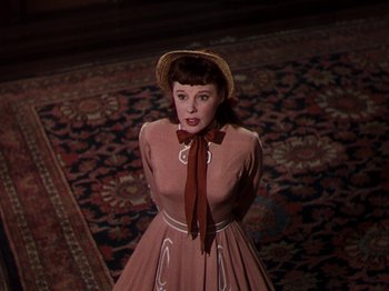 Movie still from “Little Women” (1949), directed by Mervyn LeRoy – A woman in a dress and a hat standing in front of a rug; Medium shot, High angle