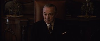 Movie still from “Live by Night” (2016), directed by Ben Affleck – An older man wearing a suit and tie sitting in a chair; Close Up shot, Low angle