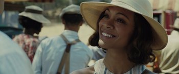 Movie still from “Live by Night” (2016), directed by Ben Affleck – A woman in a hat smiles at the camera while a man walks behind her; Close Up shot, Over the shoulder angle