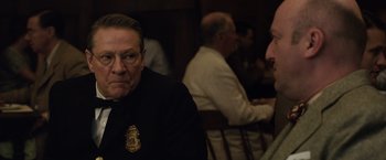 Movie still from “Live by Night” (2016), directed by Ben Affleck – An older man wearing a police uniform sitting at a table with other people; Close Up shot, Over the shoulder angle