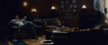 Movie still from “Live by Night” (2016), directed by Ben Affleck – A man sitting on a couch in a living room next to a table; Wide shot, High angle
