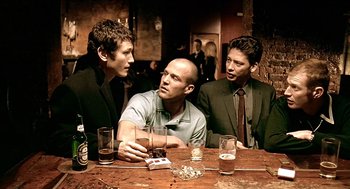 Movie still from “Lock, Stock and Two Smoking Barrels” (1998), directed by Guy Ritchie – A group of men sitting at a wooden table with drinks; Medium shot, Over the shoulder angle