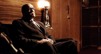 Movie still from “Lock, Stock and Two Smoking Barrels” (1998), directed by Guy Ritchie – An older man in a suit sitting in a chair in front of a lamp; Medium shot, Low angle