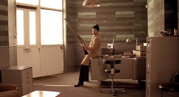 Movie still from “Lock, Stock and Two Smoking Barrels” (1998), directed by Guy Ritchie – A man sitting in front of a desk holding a rifle; Wide shot, High angle
