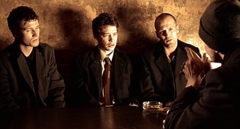 Movie still from “Lock, Stock and Two Smoking Barrels” (1998), directed by Guy Ritchie – A group of men sitting at a table in front of a brick wall; Medium shot, Low angle