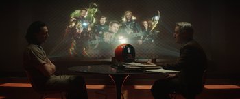 Movie still from “Loki” (2021), created by Michael Waldron – A table with a clock sitting in front of a projector screen; Wide shot, Over the shoulder angle