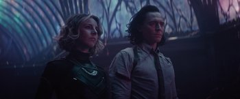 Movie still from “Loki” (2021), created by Michael Waldron – A man and a woman standing next to each other in front of trees; Medium shot, Low angle