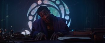 Movie still from “Loki” (2021), created by Michael Waldron – A man sitting at a table in front of a stained glass window; Medium shot, Over the shoulder angle