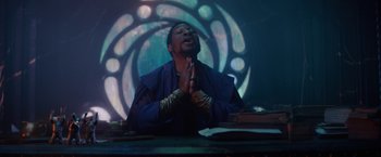 Movie still from “Loki” (2021), created by Michael Waldron – A man sitting at a table with his hands clasped in prayer; Medium shot, Low angle