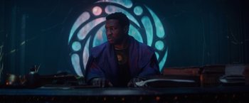 Movie still from “Loki” (2021), created by Michael Waldron – A man sitting at a table in front of a stained glass window; Medium shot, Low angle