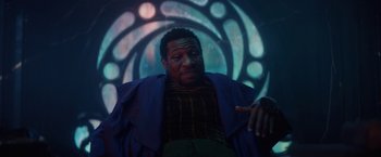 Movie still from “Loki” (2021), created by Michael Waldron – A man in a purple jacket is sitting in front of a stained glass window; Medium shot, Low angle