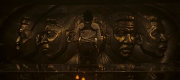Movie still from “Loki” (2021), created by Michael Waldron – A man standing in front of a wall with faces on it; Medium shot, Over the shoulder angle