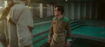 Movie still from “Loki” (2021), created by Michael Waldron – A man in a brown and green outfit standing in front of a train platform; Medium shot, Over the shoulder angle