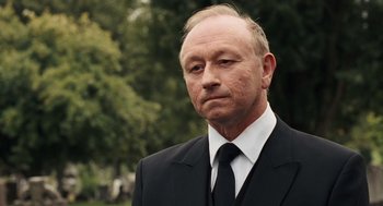 Movie still from “London Boulevard” (2010), directed by William Monahan – A man in a suit and tie standing in front of trees; Close Up shot, Over the shoulder angle
