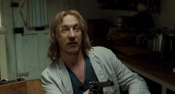 Movie still from “London Boulevard” (2010), directed by William Monahan – A man holding a gun while sitting in a room; Close Up shot, Over the shoulder angle
