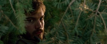 Movie still from “Lone Survivor” (2013), directed by Peter Berg – A man with a beard is looking at the camera through some bushes; Close Up shot, Over the shoulder angle