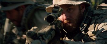 Movie still from “Lone Survivor” (2013), directed by Peter Berg – A man with a beard is holding a rifle and wearing a hat; Close Up shot, Over the shoulder angle