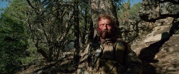 Movie still from “Lone Survivor” (2013), directed by Peter Berg – A man with blood on his face sitting next to a tree; Medium shot, Over the shoulder angle