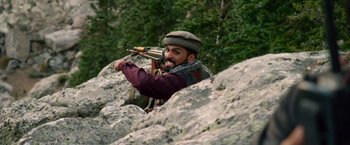 Movie still from “Lone Survivor” (2013), directed by Peter Berg – A man with a gun in his hand is standing on a rocky cliff; Medium shot, Over the shoulder angle