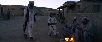 Movie still from “Lone Survivor” (2013), directed by Peter Berg – A man standing next to a fire in the middle of a dirt field; Wide shot, Over the shoulder angle