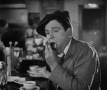Movie still from “Lonesome” (1928), directed by Pál Fejös – An old photo of a man sitting at a table eating a donut; Medium shot, Low angle