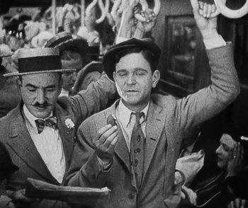 Movie still from “Lonesome” (1928), directed by Pál Fejös – A man in a suit and hat is holding his hands in the air while another man in a suit and hat is holding his hands up; Medium shot, High angle