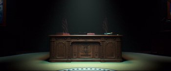 Movie still from “Long Shot” (2019), directed by Jonathan Levine – A large wooden desk in front of two american flags; Wide shot, High angle