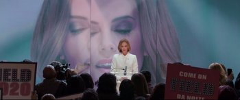 Movie still from “Long Shot” (2019), directed by Jonathan Levine – A woman in a white suit standing in front of a microphone; Medium shot, Over the shoulder angle