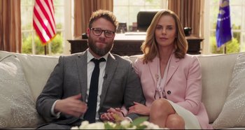 Movie still from “Long Shot” (2019), directed by Jonathan Levine – A man and a woman sitting next to each other on top of a couch; Medium shot, Over the shoulder angle