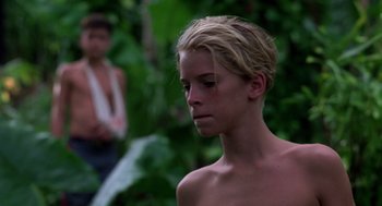 Movie still from “Lord of the Flies” (1990), directed by Harry Hook – A young man with blonde long hair and no shirt is standing in the woods; Close Up shot, Low angle