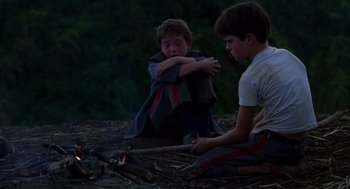 Movie still from “Lord of the Flies” (1990), directed by Harry Hook – Two young boys sitting on the ground near a fire; Medium shot, Over the shoulder angle