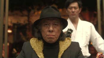 Movie still from “Love Exposure” (2008), directed by Sion Sono – An older man wearing a black hat and a black jacket; Close Up shot, Low angle