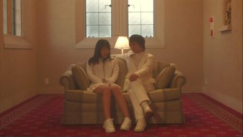 Movie still from “Love Exposure” (2008), directed by Sion Sono – A man and a woman sitting on a couch in front of a window; Wide shot, Low angle