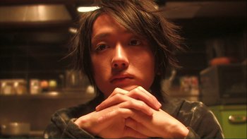 Movie still from “Love Exposure” (2008), directed by Sion Sono – A man with long black hair is posing for a picture; Close Up shot, Low angle