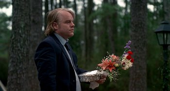 Movie still from “Love Liza” (2002), directed by Todd Louiso – A man in a suit holding a tray of flowers in front of a wooded area; Medium shot, Over the shoulder angle