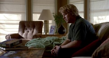 Movie still from “Love Liza” (2002), directed by Todd Louiso – A man sitting in a living room next to a table; Medium shot, Over the shoulder angle